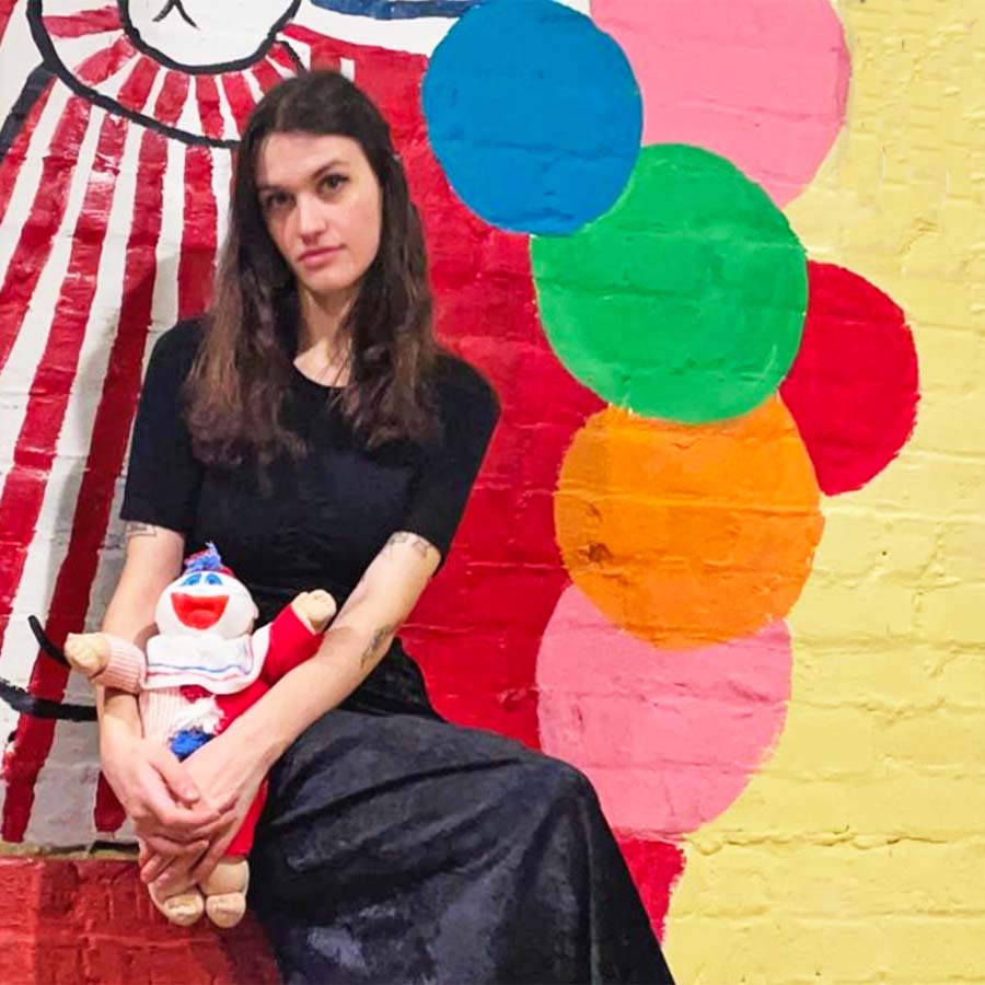 Chloe Manon sits somberly in front of a brightly painted wall within Graveface Museum, holding a freaky Pogo the Clown doll.