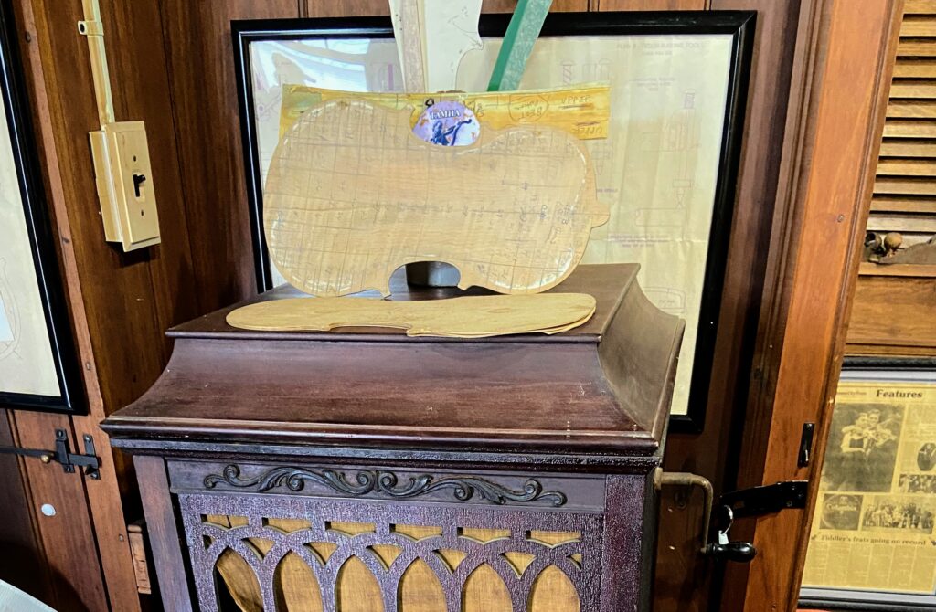 The image depicts an unfinished violin body displayed on a wooden phonograph inside the Ralph Blizzard museum. Behind the violin, several documents, newspaper clippings, and a blueprint are framed, detailing violin construction and related musical notes. The overall setting is a historical exhibit, with rich wood paneling adding to the ambiance.