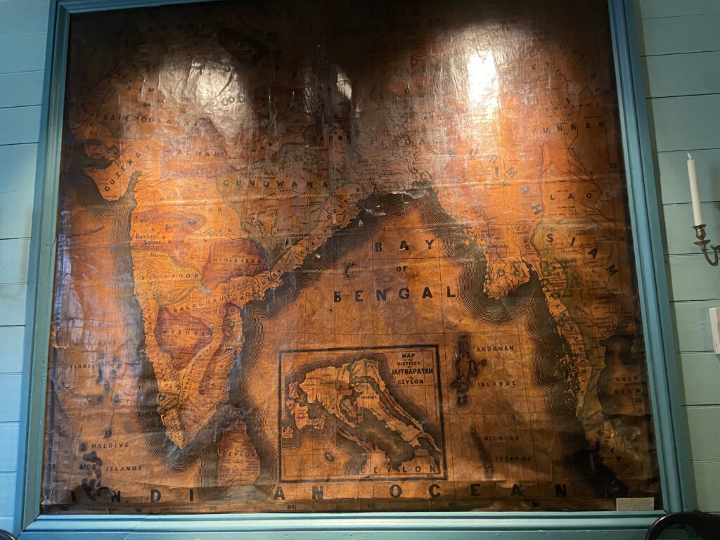 The image displays a large, vintage map of the Indian subcontinent framed and mounted on a wall. The map, which appears aged and worn, features detailed geographical labels including major cities like Bombay and Calcutta, regions such as Ceylon and Mysore, and bodies of water like the Bay of Bengal. The map is illuminated by overhead lighting, casting a warm glow over the textured surface.
