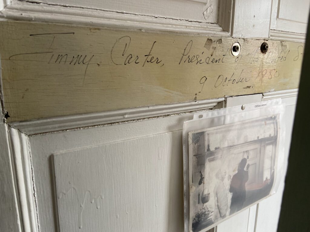 The image shows a close-up view of a worn door frame with the hand-written inscription "Jimmy Carter, President of the U.S.A., 9 October 1980." Below this inscription, there is a small, clear plastic pocket attached to the door containing a faded black and white photograph of two people. The entire scene is tinged with a vintage, historical ambiance.