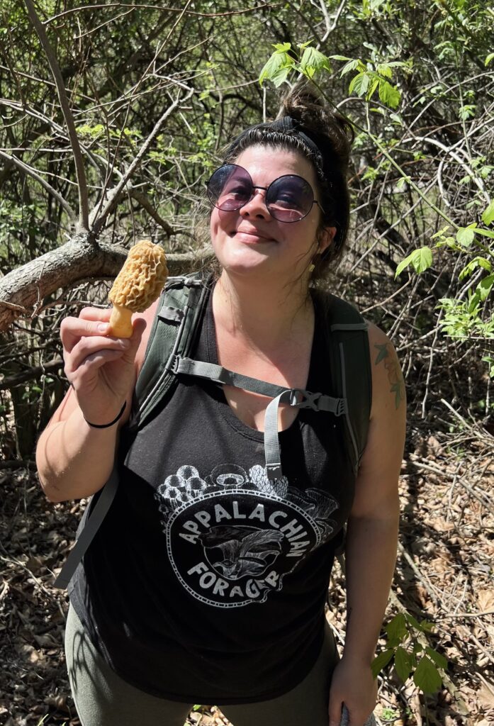 Whitney Johnson, known as the Appalachian Forager, smiles while holding a morel mushroom in a forested area. She is wearing circular sunglasses and a black tank top with the 'Appalachian Forager' logo, and has a backpack on, surrounded by green foliage and trees.