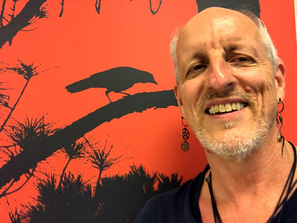 Rick de Yampert, also known as Mr. Crow, smiling in front of his artwork. The background features a vibrant orange canvas with a black silhouette of a crow perched on a branch amidst pine trees