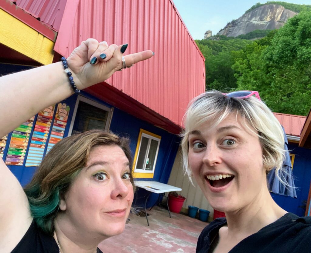 Becky and Diana, hosts of the Homespun Haints podcast, share a laugh while pointing at Chimney Rock in North Carolina, poking fun at its amusing weenie shape. Their mischievous expressions add a lighthearted touch to their adventurous paranormal Appalachian road trip.