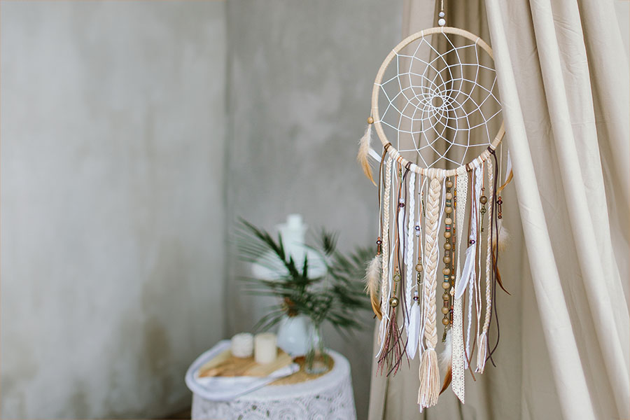 A dreamcatcher in neutral colors made from a hoop, feathers, and beads hangs inside a window to protect the home's occupants from bad dreams and bad energy while they sleep.