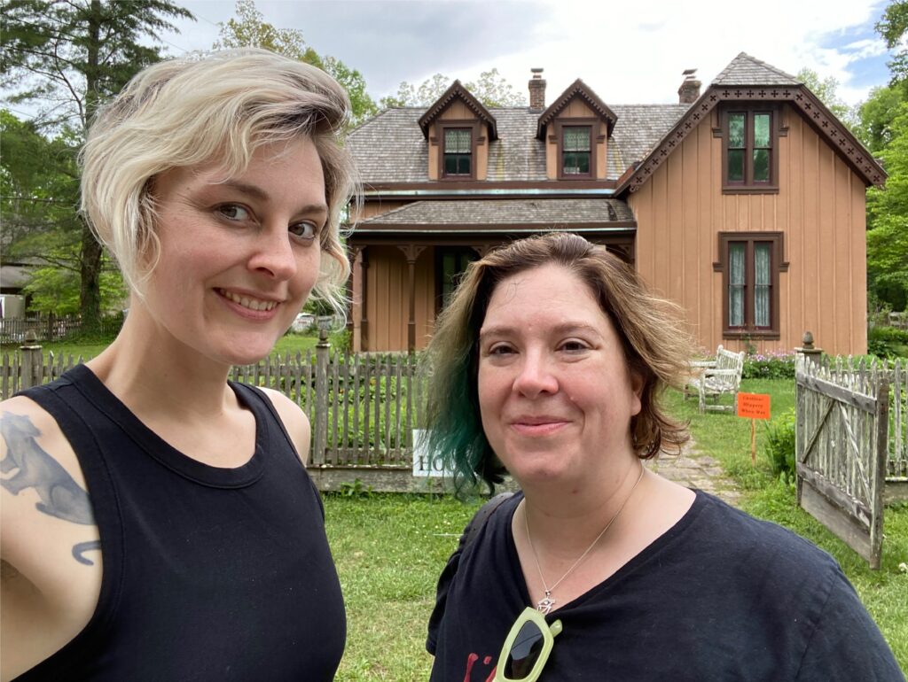 Diana and Becky of Homespun Haints podcast, smiling in front of Madame Hughes' house at the historic Town of Rugby, a picturesque and haunted site on their Appalachian road trip, with its charming Victorian architecture and picket fence in the background.