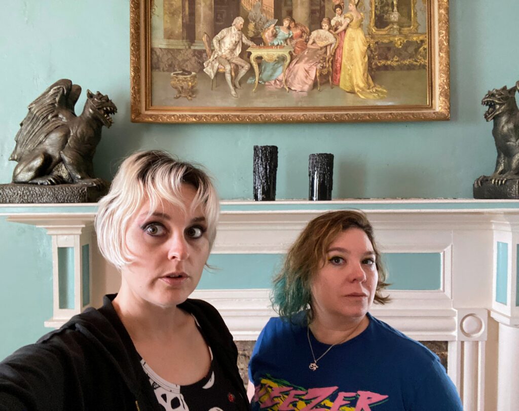 Becky and Diana, hosts of the Homespun Haints podcast, standing solemnly in front of the white and aqua carved fireplace mantle adorned with spooky gargoyles, dripping black pillar candles, and a gilt-framed Rococo painting of young women in silk gowns, at the haunted Nickerson-Sneed House.