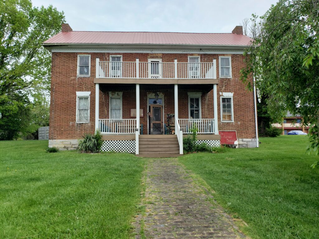 Diana, co-host of the Homespun Haints podcast, peeking out of of the historic and haunted Nickerson Sneed House in Glade Spring, VA, a notable site on a paranormal Appalachian road trip, showcasing the classic brick architecture surrounded by lush greenery.