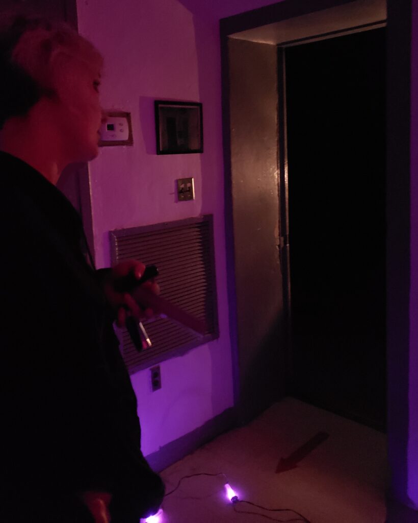 Diana, co-host of Homespun Haints podcast, using a new ghost hunting tool called Trip Wire (which is like a battery powered string of violet lights that change color in response to localized EMF spikes) during a paranormal investigation at the haunted Old Scott County Jail. The image captures a moment with Diana illuminated by the purple light of the Trip Wire in a dimly lit hallway outside of a second story jail cell, enhancing the eerie atmosphere.