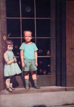 The image shows a boy and a girl standing in front of a window. The boy, in a blue shirt and shorts, stands to the right, while the girl, wearing a blue dress, stands to the left. The window behind them is dark, with hands pressed against the glass, adding an eerie atmosphere to the scene.