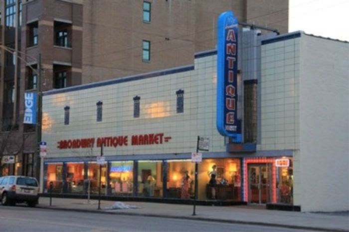 The image shows the exterior of a building named "Broadway Antique Market." The facade has a retro design with large windows displaying various antiques, and a prominent vertical sign that reads "Antique" in neon letters. The scene is set in an urban area with neighboring buildings visible, and a parked car is seen in front. The storefront is brightly lit, giving it a welcoming appearance, despite the possibility that some of the antiques within the shop might be haunted.