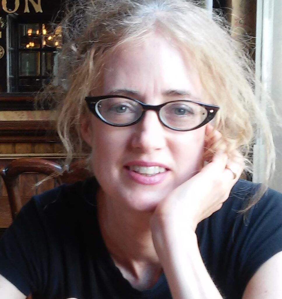 Headshot of Beth Castrodale, author of 'The Inhabitants.' She has light blonde hair tied back messily, wears black-rimmed glasses, and is smiling softly while resting her chin on her hand. The background suggests a cozy indoor setting with warm lighting, very different from the architecture of the haunted houses we talk about in today's episode.