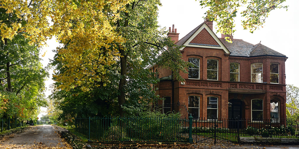 A large, old brick house with tall, narrow windows and a steeply pitched roof, surrounded by tall trees with autumn foliage. The gothic architecture features pointed gables, intricate brickwork, and a slightly overgrown garden, giving the structure the classic features of haunted houses. The scene is set in daylight, but the house's dark windows and shadowy corners suggest a mysterious, possibly eerie atmosphere.