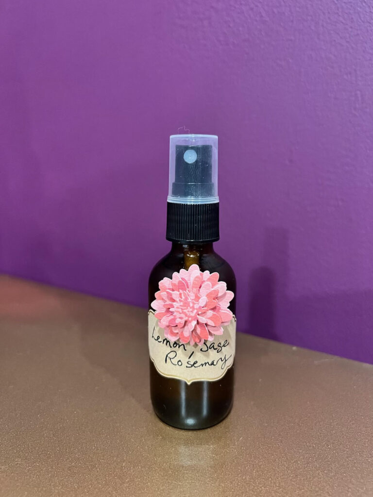 A lovely brown glass bottle with a pink chrysanthemum sticker, containing a homemade concoction we call Becky and Diana's Lazy Haint Spray, possibly ideal for clearing out ghosts at the Fall Equinox.