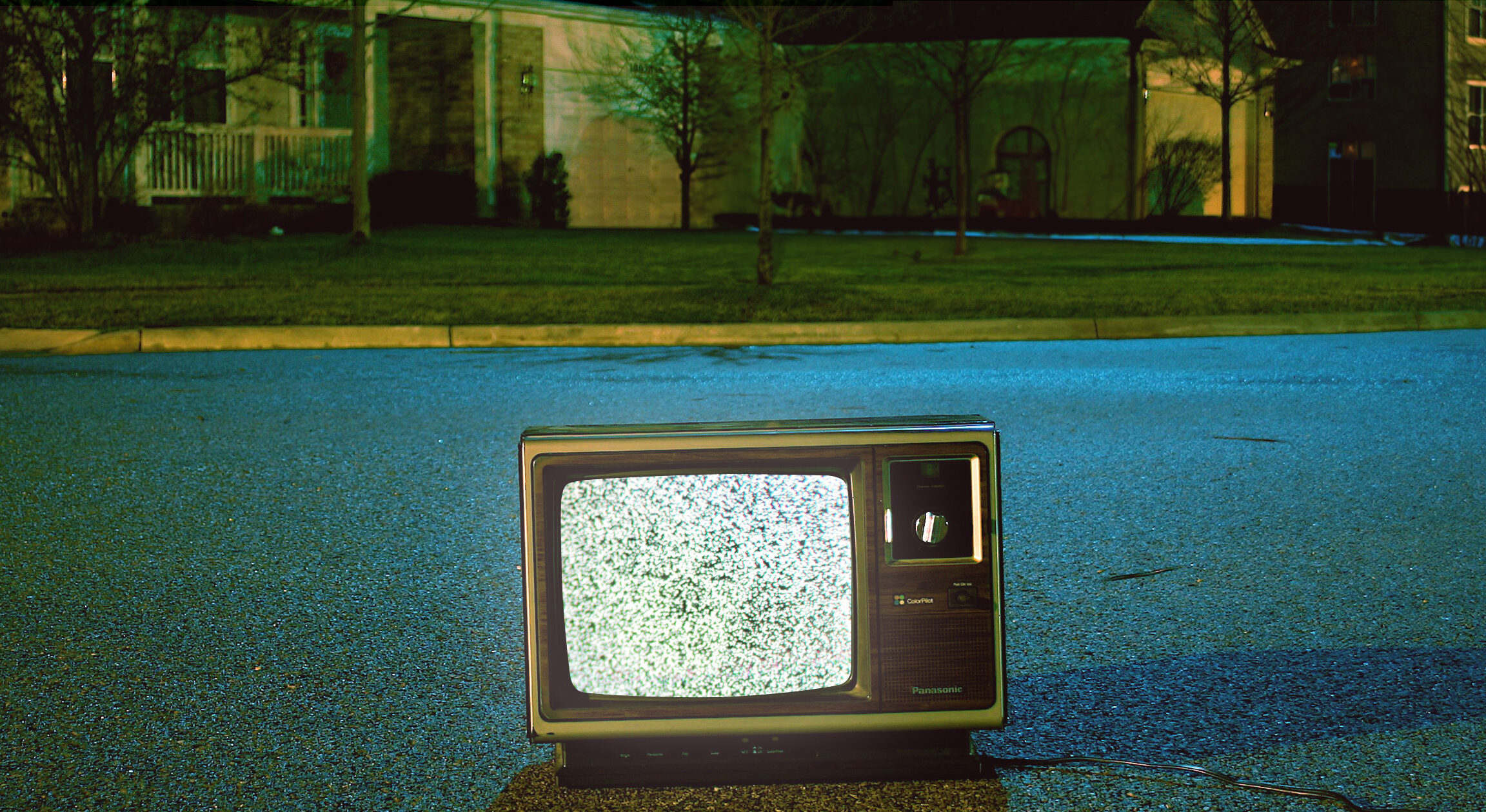 A vintage television with a static-filled screen sits in the middle of a quiet suburban street at night, with its power cord trailing behind. In the background, houses with dim lighting are visible under a deep blue sky, creating a spooky and surreal atmosphere.