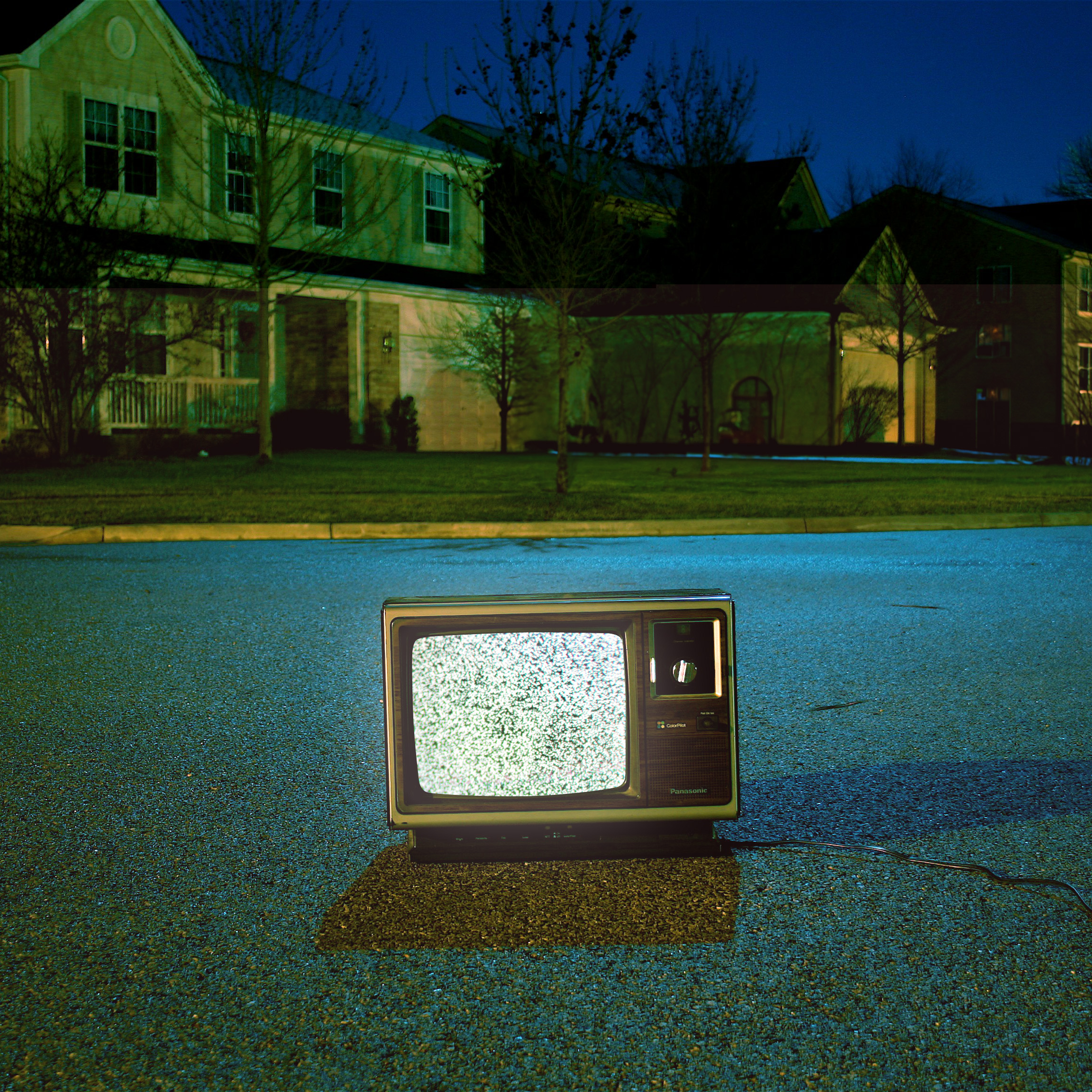 A vintage television with a static-filled screen sits in the middle of a quiet suburban street at night, with its power cord trailing behind. In the background, houses with dim lighting are visible under a deep blue sky, creating a spooky and surreal atmosphere.