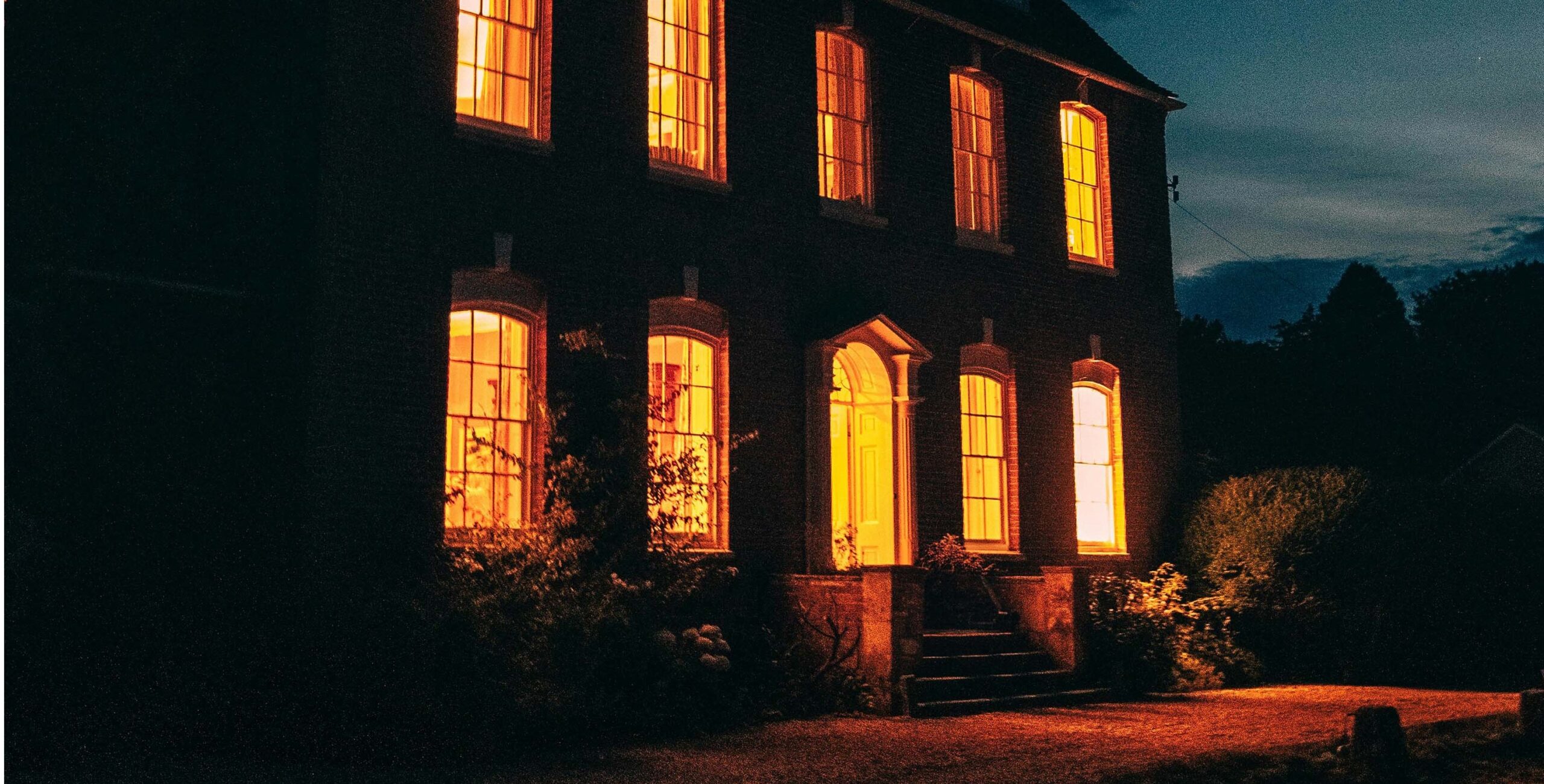 This image shows a large southern gothic mansion at night with warm yellow-orange lights glowing from its windows, creating an eerie, mysterious ambiance. The house is an interpretive rendering of the Mississippi mansion where Stacey and friends held their séance using her Southern Gothic Oracle deck. The edifice stands in contrast to the dimly lit surroundings and dark sky, evoking a spooky feel.