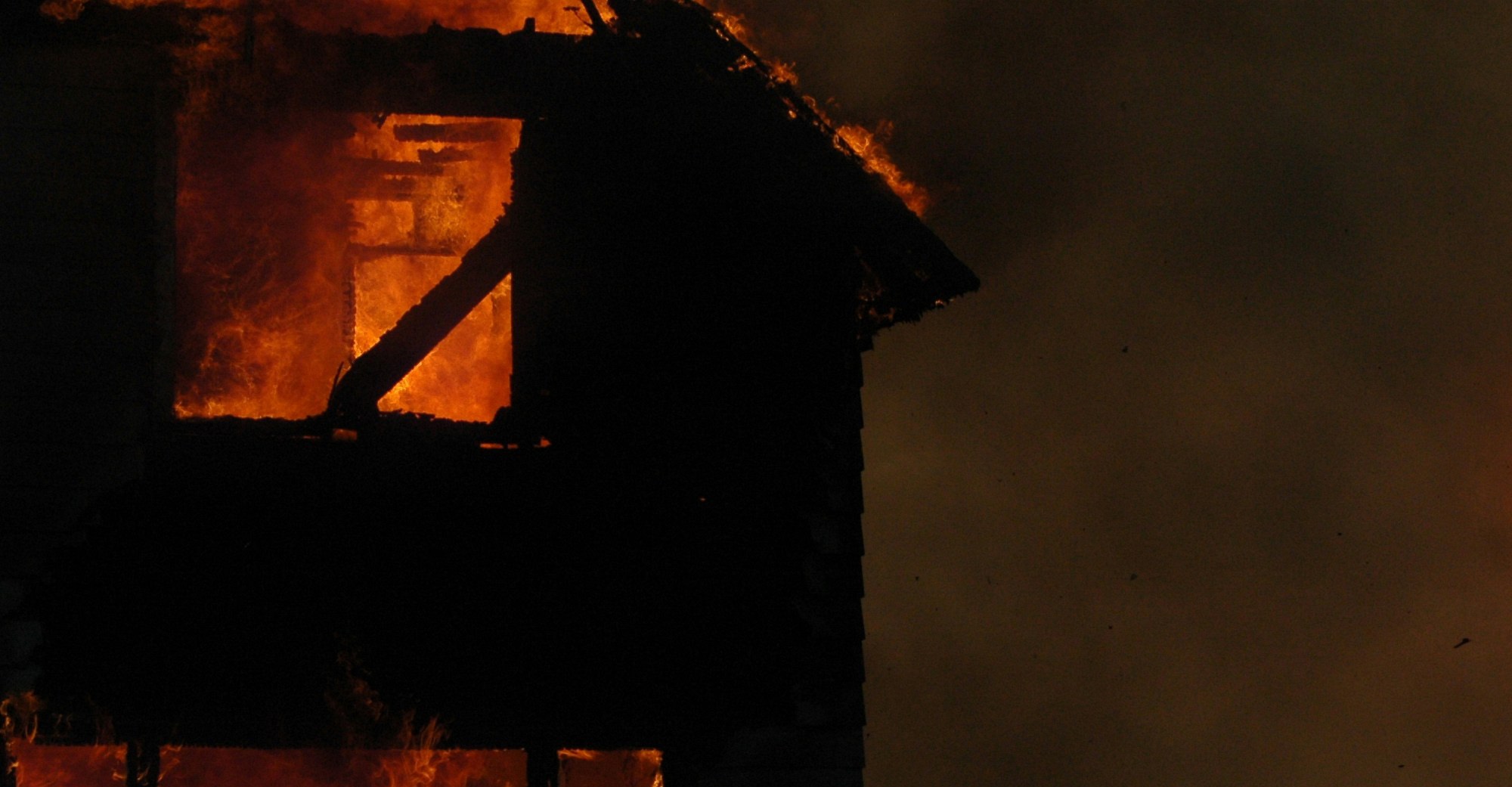 A house fire burns brightly in the dark of night, consuming both stories of the house pictured. This represents the house fire Ashely experienced in childhood that convinced him ghosts can hurt you.