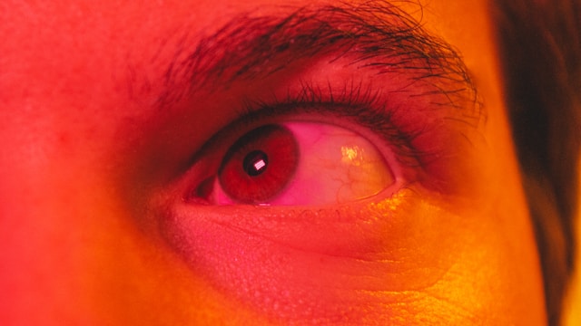 This close-up image captures a person's eye bathed in an intense red and orange light, creating a dramatic and slightly unsettling effect. The red hue gives the eye a possessed appearance, like someone who has awoken to experience sleep paralysis. The sharp detail on the eyelashes and iris adds to the striking, almost hypnotic quality of the image.