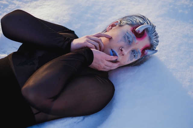 This image shows an androgynous person lying on snow with a surreal, artistic makeup look that includes small red horns, blue lipstick, and intricate white designs around the eyes, creating an icy, otherworldly appearance. They wear a black, semi-sheer top, contrasting starkly against the snowy background. The expression and styling evoke a mystical, wintery creature, blending elements of fantasy and cold beauty.