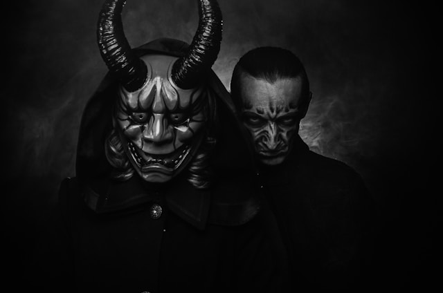 This black-and-white image features two figures in eerie, theatrical masks set against a dark, smoky background. The foreground figure wears a large, menacing horned mask with sharp teeth, exuding a demonic or oni-like appearance, while the figure behind has a subtler, sinister mask with intense, shadowed features. The composition and lighting create a haunting, ominous atmosphere, emphasizing themes of horror and the supernatural.