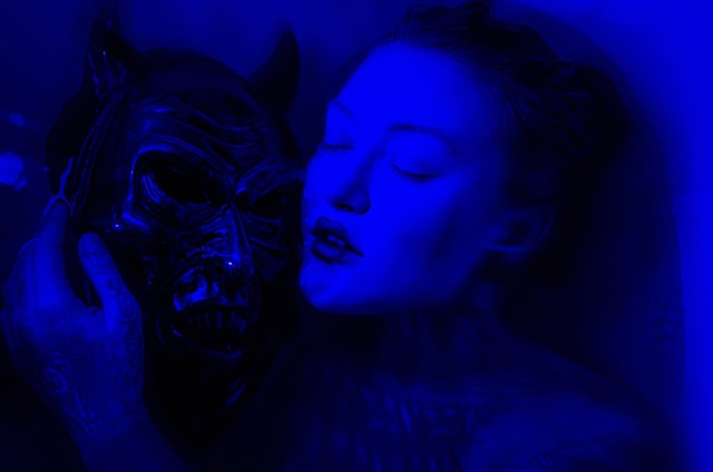 This image, bathed in an intense blue light, shows a feminine person resting with eyes closed, embracing their sleep paralysis demon with a gentle caress. The contrast between the human and the menacing dark demon creates an intriguing juxtaposition, blending elements of macabre allure. 