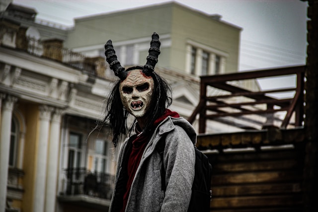 The image depicts a person wearing a frightening, demon-like mask with twisted horns, sharp fangs, and hollow eyes, standing outdoors against a backdrop of old European-style buildings. The individual is dressed in a gray coat and appears to be in an urban setting, creating a juxtaposition between the eerie mask and the everyday environment. The dark, dramatic style adds an unsettling, horror-inspired atmosphere to the scene.