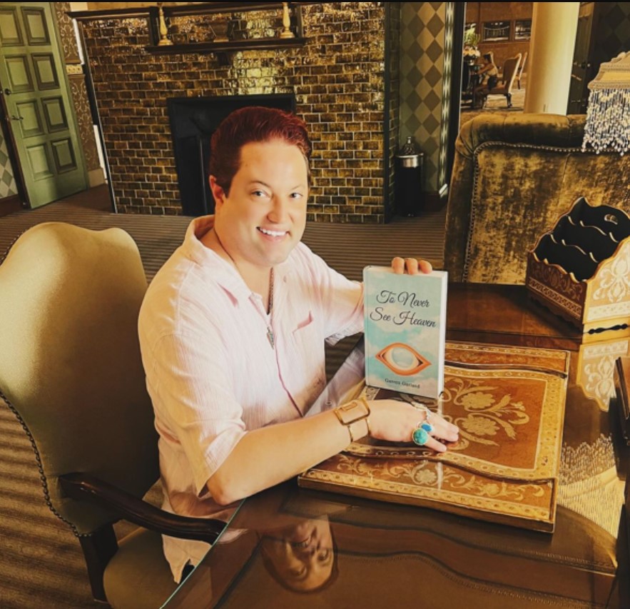 Garrett Garland, the author of 'To Never See Heaven,' smiles while seated at an ornate wooden table, holding up a copy of his book. The room features a cozy vintage interior with a fireplace and decorative furnishings, adding warmth to the setting.