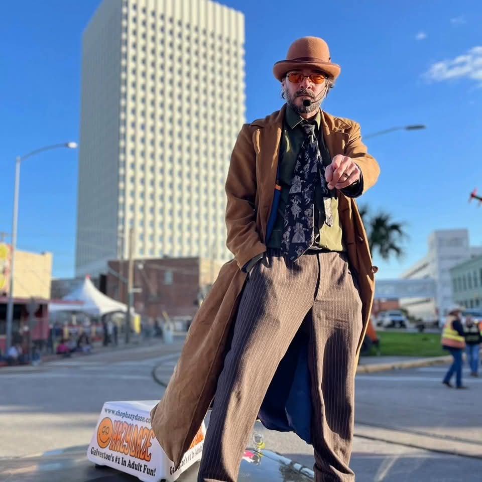 Galveston Texas tour guide Eddy Specter resembles a unique iteration of The Doctor, looking dapper in his brimmed hat and orange-lensed glasses, points directly at the camera as his camel colored duster billows majestically behind him. In the background, a skyscraper and a street festival tent are out of focus. He might just be standing on top of a vehicle.