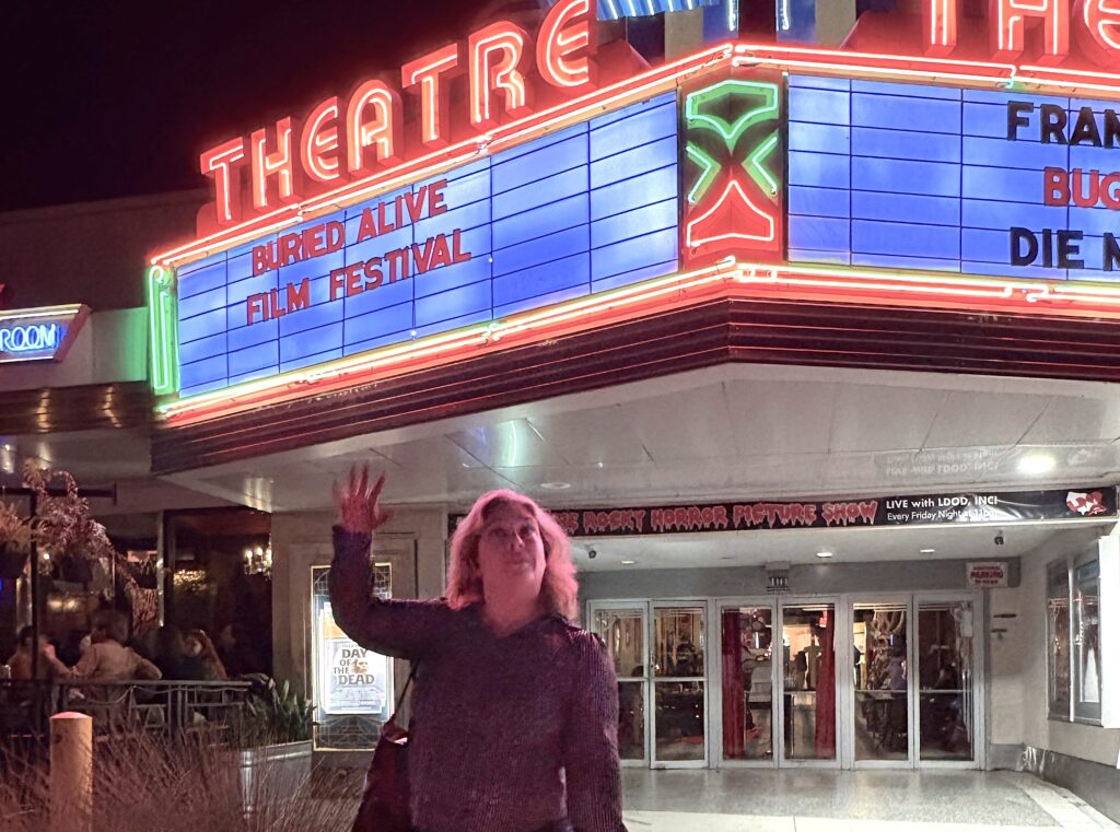 Becky of Homespun Haints poses with a wave of her hand in front of the bright red neon marquee at the Plaza Theater Atlanta, which announces in red letters "Buried Alive Film Festival"